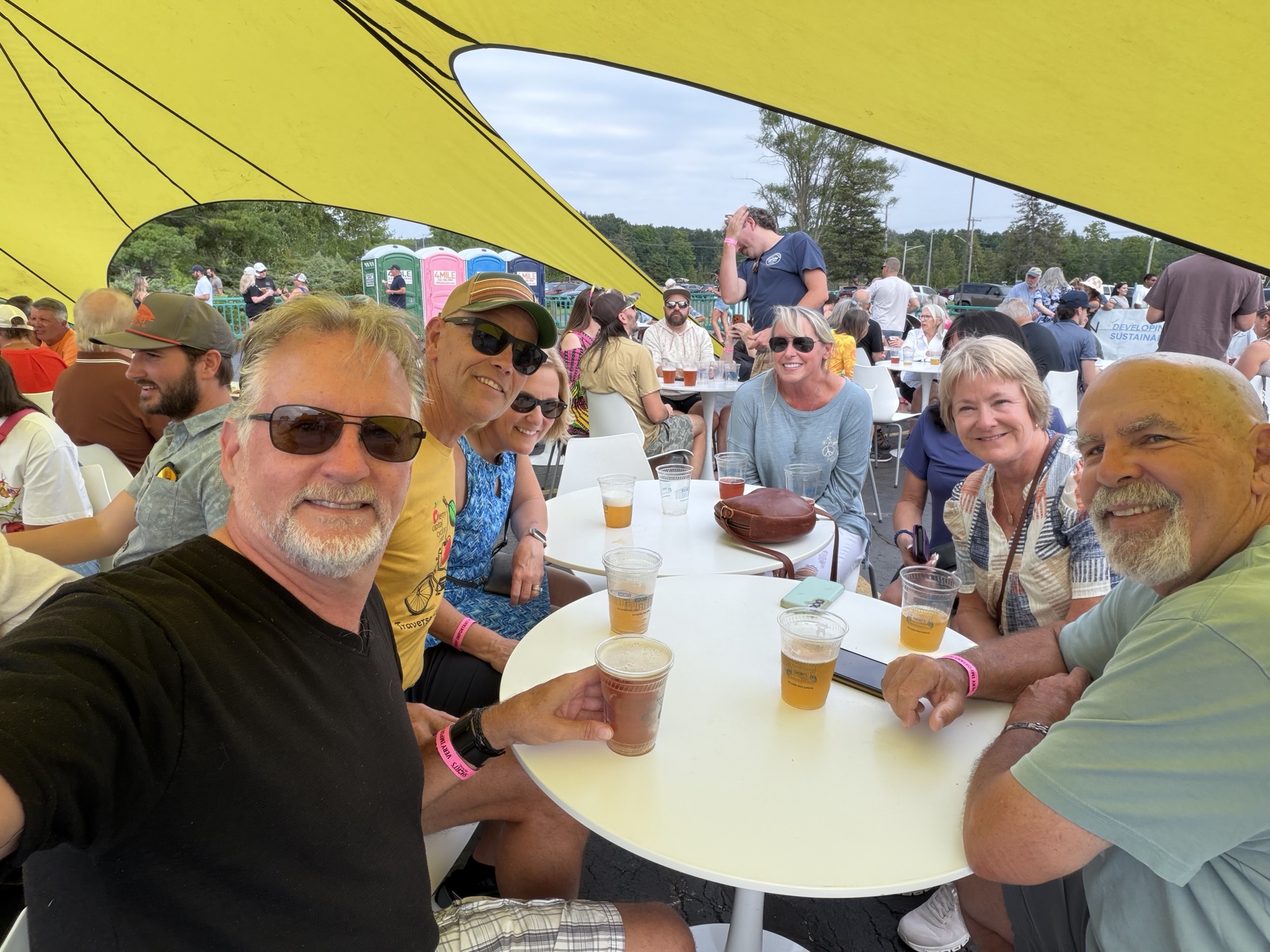 Group of friends enjoying drinks at a table under a yellow tent, smiling and socializing at an outdoor event.