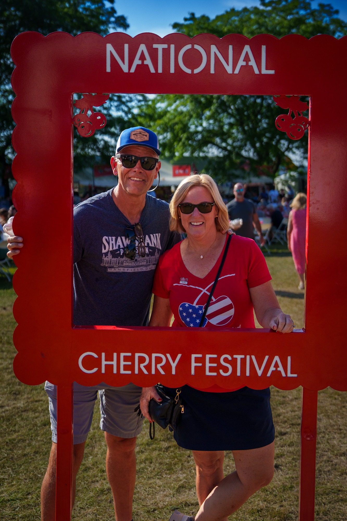 A couple poses together in a red decorative frame that reads 'NATIONAL CHERRY FESTIVAL', with a festive background of people enjoying the event.